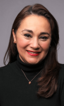 Retrato de una mujer sonriendo, con fondo gris neutro. Lleva un suéter negro de cuello alto, pendientes dorados y un collar delicado. Su cabello es largo y castaño, peinado hacia un lado.