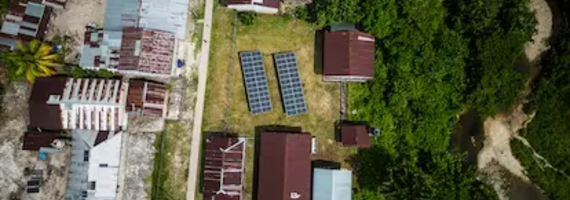 Top-down aerial view of two large solar panel arrays installed in a grassy area between small houses and a forest