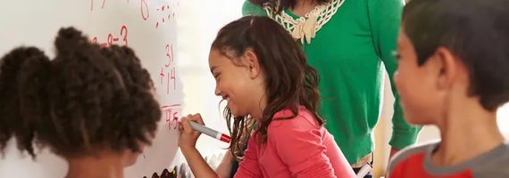 A young girl smiling while writing math equations on a whiteboard during a classroom lesson with classmates and a teacher