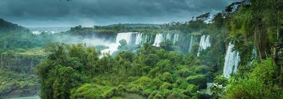 Wide view of multiple massive waterfalls cascading over lush green cliffs in a tropical rainforest under a cloudy sky