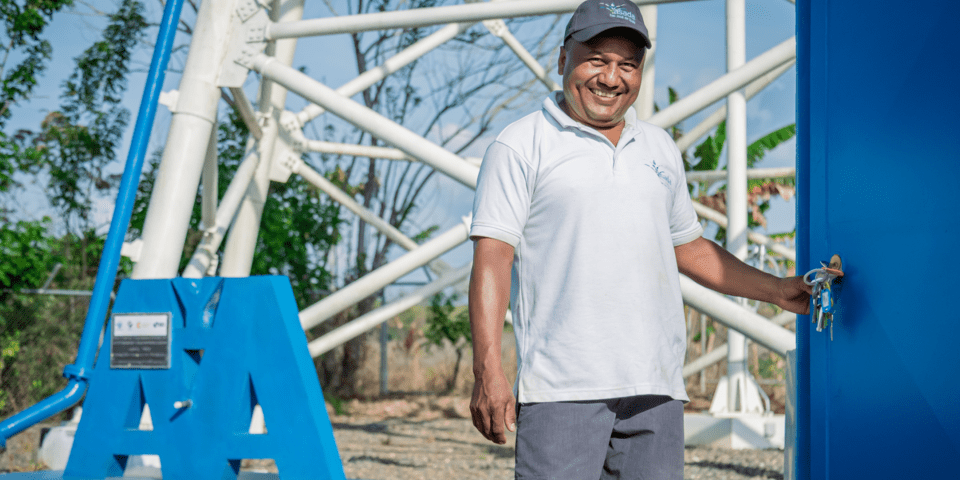 Dark-skinned man standing smiling and opening a door with a structure in the background - Inter American Development Bank - Sustainable Development