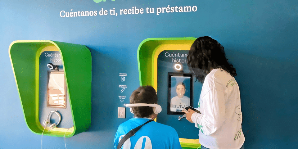Man sitting with his back to the camera wearing headphones and woman standing pointing at a touchscreen - Inter American Development Bank - IDB Lab