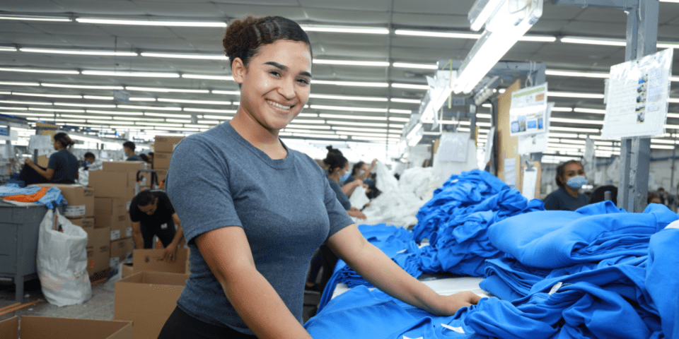 Woman smiling with a clothing factory in the background - Inter American Development Bank - Impact Evaluation