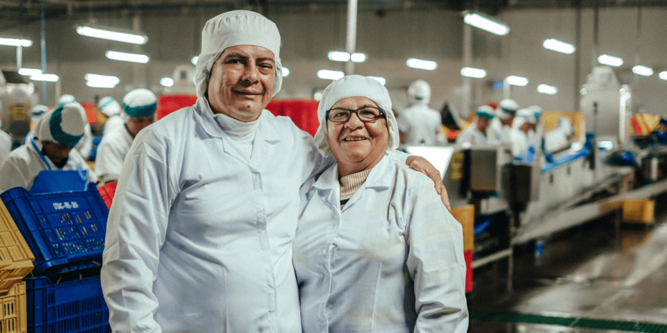 Elderly man and woman smiling and standing inside a factory with more workers and machines in the background - Inter American Development Bank - Labor Markets