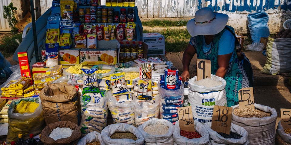 Woman in a wide-brimmed hat arranges products at an open-air market stall, with sacks of grains and cereals and packaged food items on display, prices handwritten on cardboard signs.