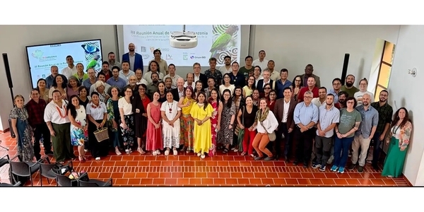 Participants of the III Annual Meeting of the Bioamazonia Network are centered in the image, posing for the event’s official photo. In the background, there is a white wall displaying the projection of the event banner.
