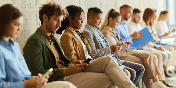 Diverse group of job candidates seated in a row, some looking at their phones and others holding application materials.