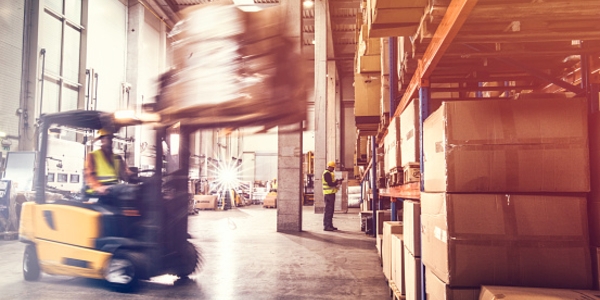 A forklift transporting a pallet of goods through a warehouse, with a worker standing near storage shelves.