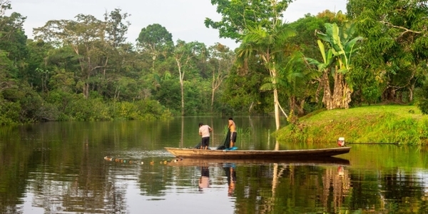 Two people fish from a wooden boat on a calm river surrounded by dense Amazonian vegetation.