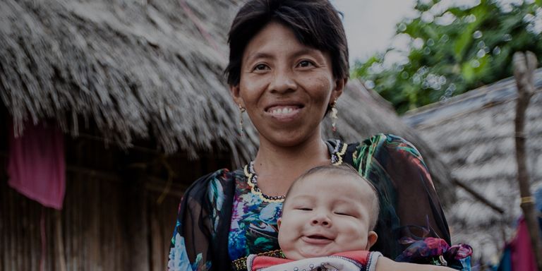 An indigenous woman is carrying her baby in front of a hut - Inter-American Development Bank - IDB