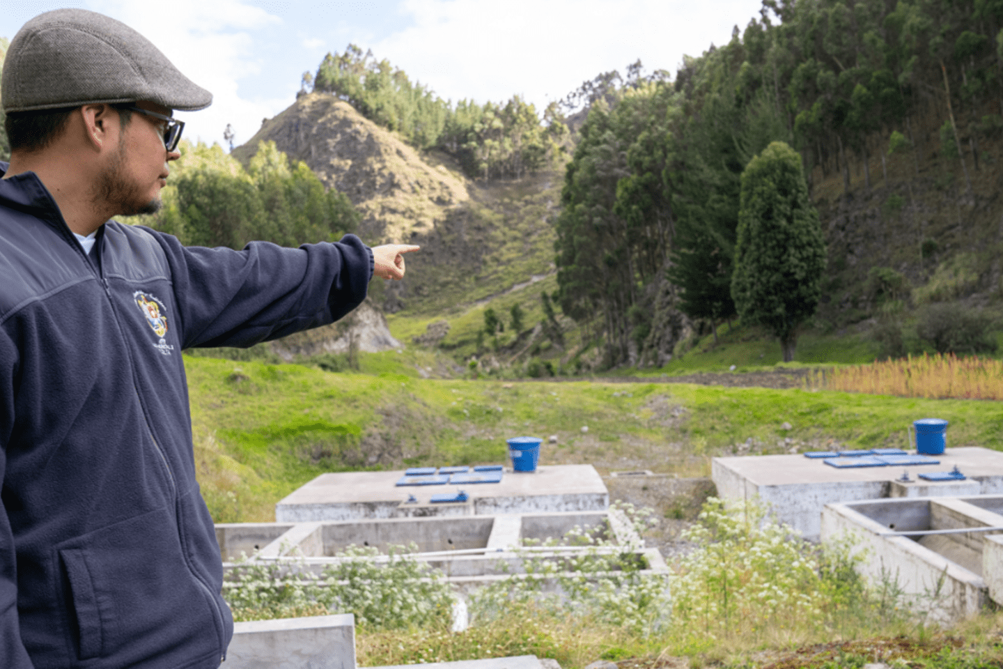 Hombre con gorra plana y chaqueta de forro polar gris está de pie en un campo verde y estructuras de hormigón  sistema de tratamiento de agua 