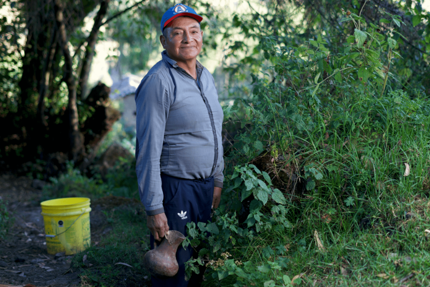 Hombre sonriente con gorra, una camisa gris y pantalones oscuros, se encuentra en la Pampa Hermosa en Perú sostiene un jarón con agua