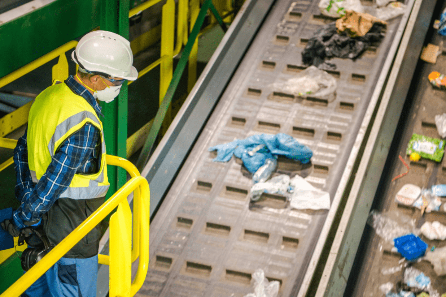 Trabajador con casco blanco, chaleco amarillo y mascarilla mirando  plataforma para impulsar la economía circular y reducir las emisiones  llena de residuos y materiales reciclables 