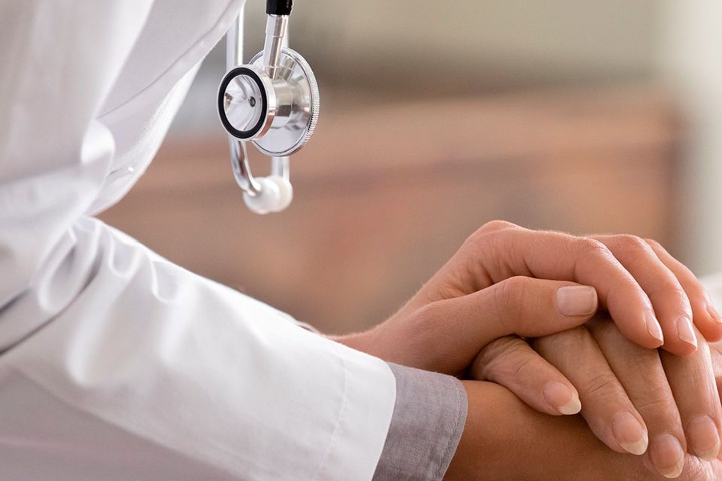 Person in a doctor's coat and with a stethoscope holding hands with a smiling woman, likely to be a patient.