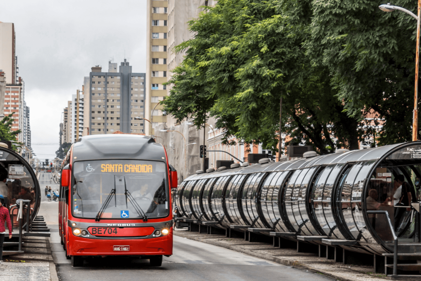 Bus rojo del sistema de transporte llegando a una estación tipo tubo en una avenida urbana, con personas esperando y edificios residenciales al fondo.