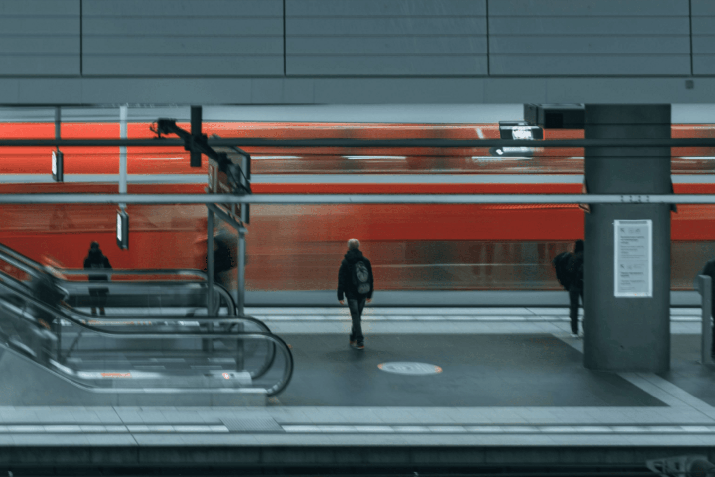 Andén de una estación de tren subterránea donde un tren rojo pasa rápidamente. Varias personas esperan o caminan por la plataforma, y se observan escaleras mecánicas y señales dentro del espacio moderno.