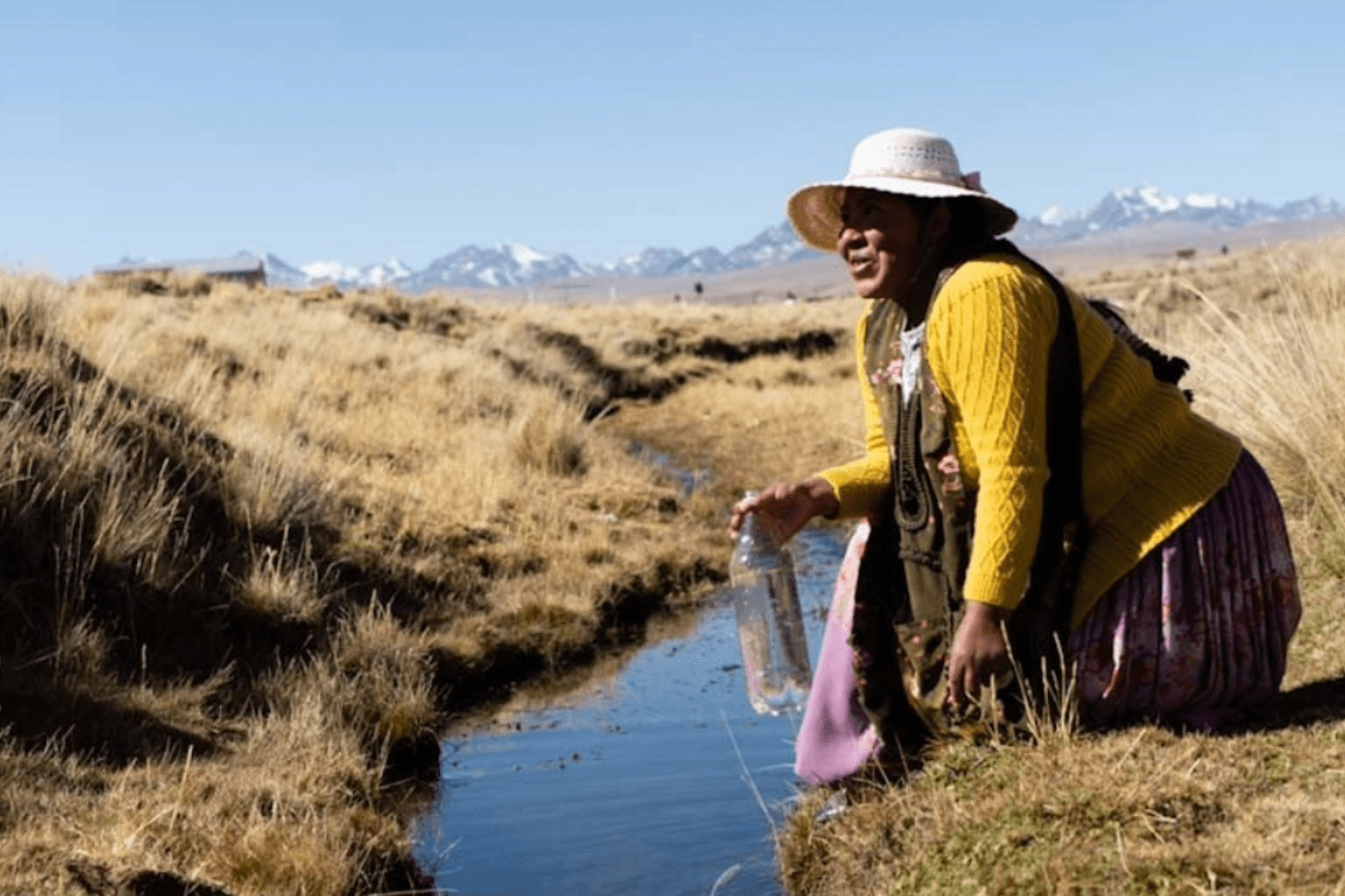 A rural woman wearing a hat and traditional clothing kneels beside a small stream of water and sanitation in a grassy, mountainous landscape, holding a plastic bottle.