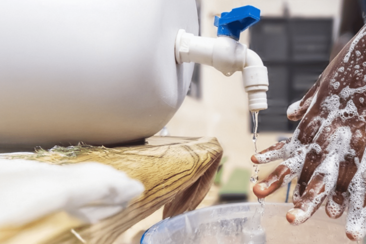 A person outdoors practicing hand hygiene using a simple handwashing setup consisting of a plastic water container and a bucket.