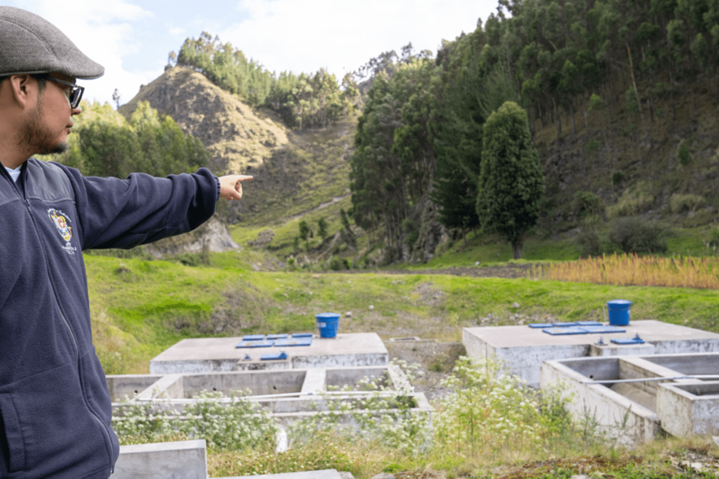 A man wearing a gray cap, glasses, and a dark jacket is pointing to concrete structures in a rural, hilly landscape with water that transforms lives in rural communities in Ecuador.
