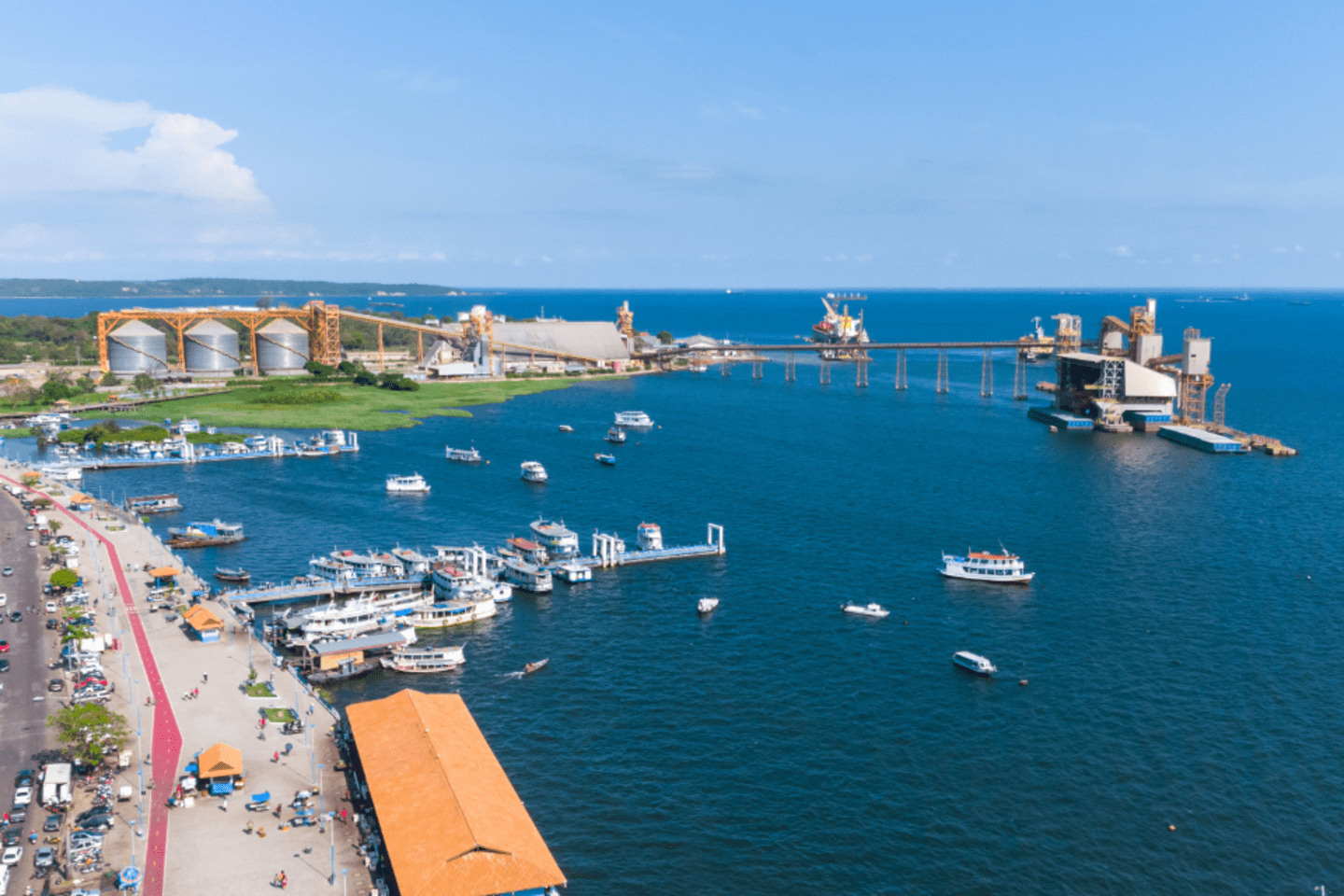 Vista aérea de un puerto fluvial con varios barcos y lanchas atracados en aguas azules. A la izquierda se observa una avenida con tráfico y edificios, mientras que al fondo hay instalaciones industriales con grandes silos y estructuras sobre el agua. El cielo está despejado con algunas nubes en el horizonte.