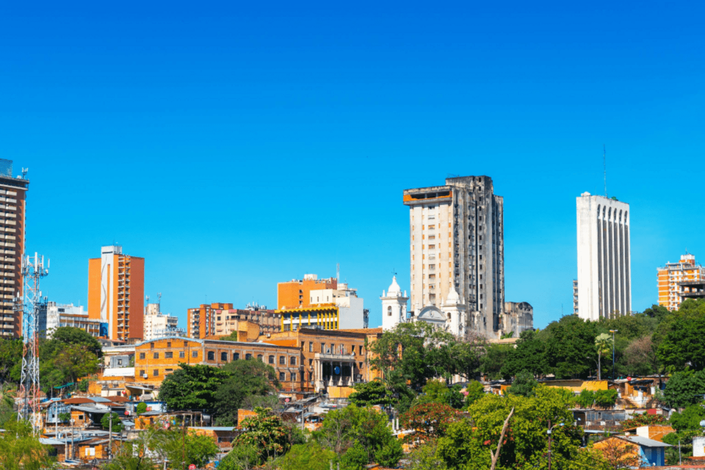 Vista panorámica de una ciudad con edificios altos y modernos mezclados con construcciones más antiguas y áreas verdes en primer plano. El cielo está despejado y de color azul intenso, destacando las torres y estructuras urbanas