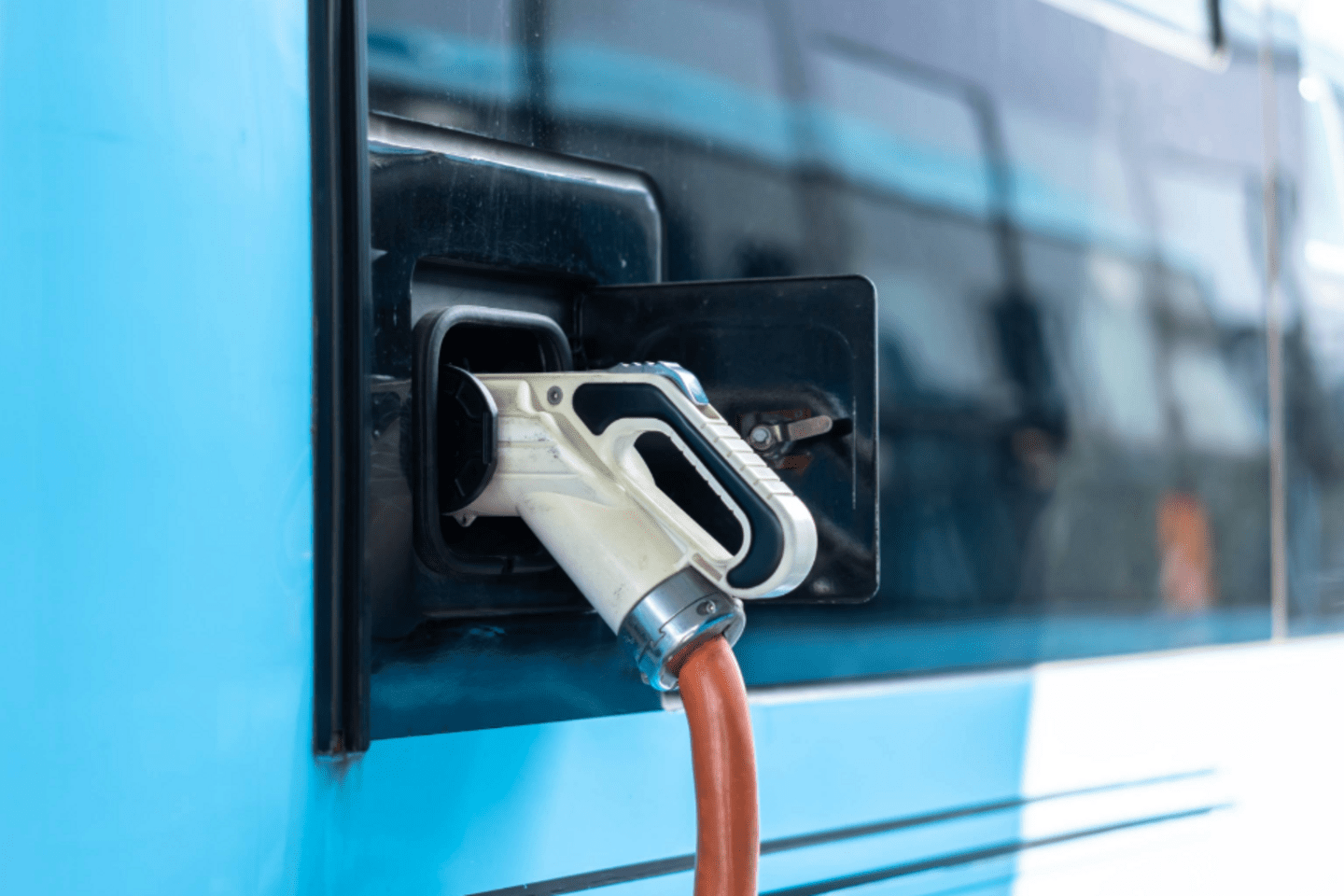A close-up view of an electric vehicle charging port with a charger plugged in. The vehicle is light blue, and the charging cable is orange, connected securely to the side of the vehicle.