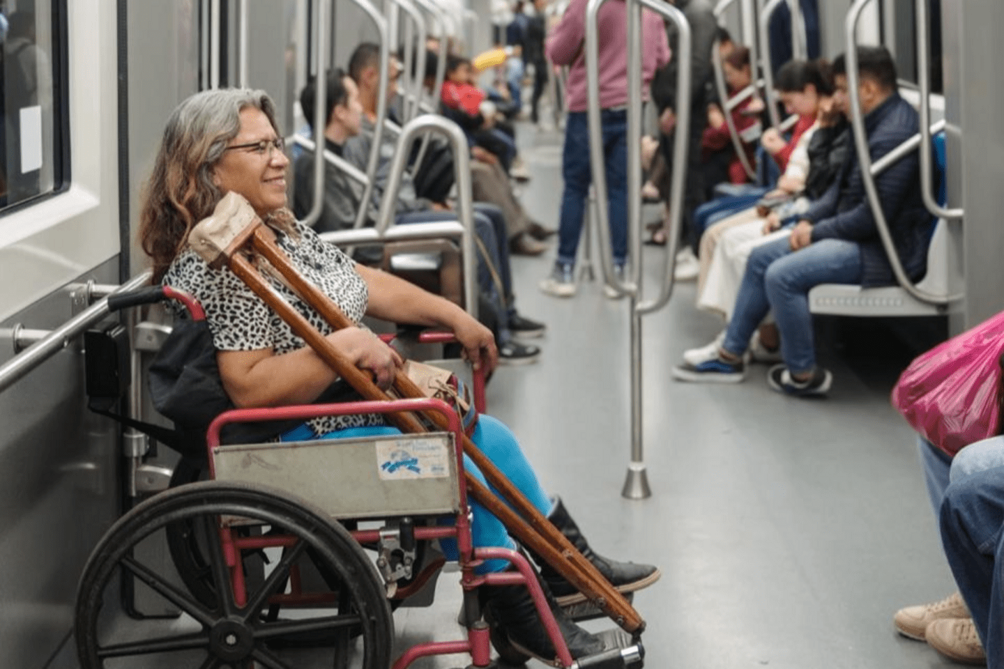 Woman using a wheelchair and holding crutches while riding on a metro train, with other passengers seated along the aisle.