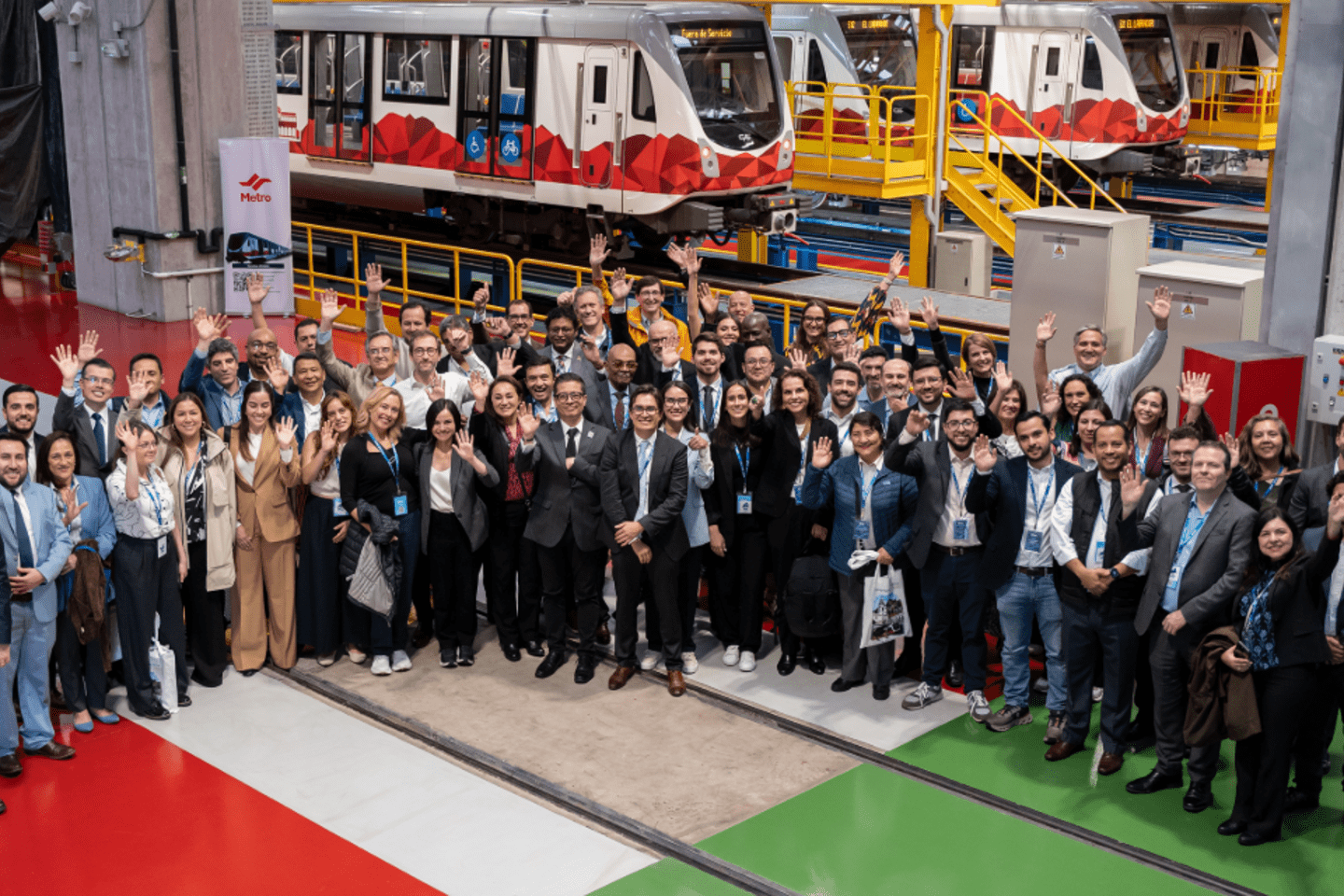 Grupo numeroso de personas posando para una foto en el interior de una instalación industrial, con varios trenes del metro estacionados en el fondo sobre vías elevadas y estructuras amarillas. El suelo es de colores rojo, verde y gris, y se observan columnas y equipos alrededor