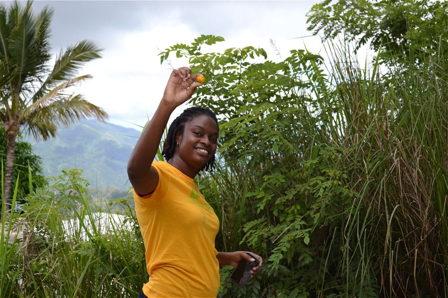 A young woman in Jamaica holds up her hand to show a fruit, the result of a projects protecting watersheds from degradation, preserving their ecological integrity, and the key resource they provide.