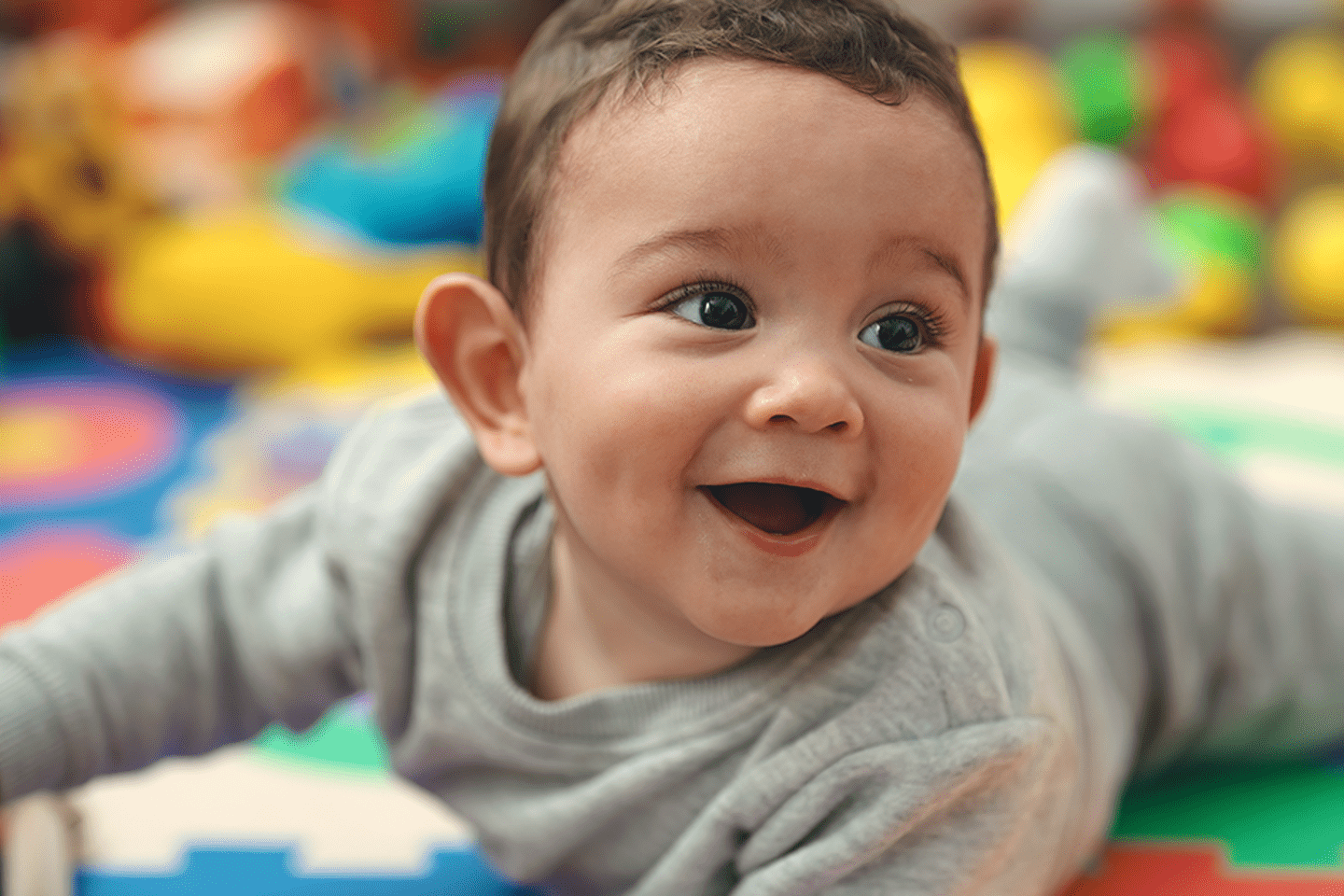 A smiling baby lies on a colorful playmat, looking to the side in a light gray outfit.