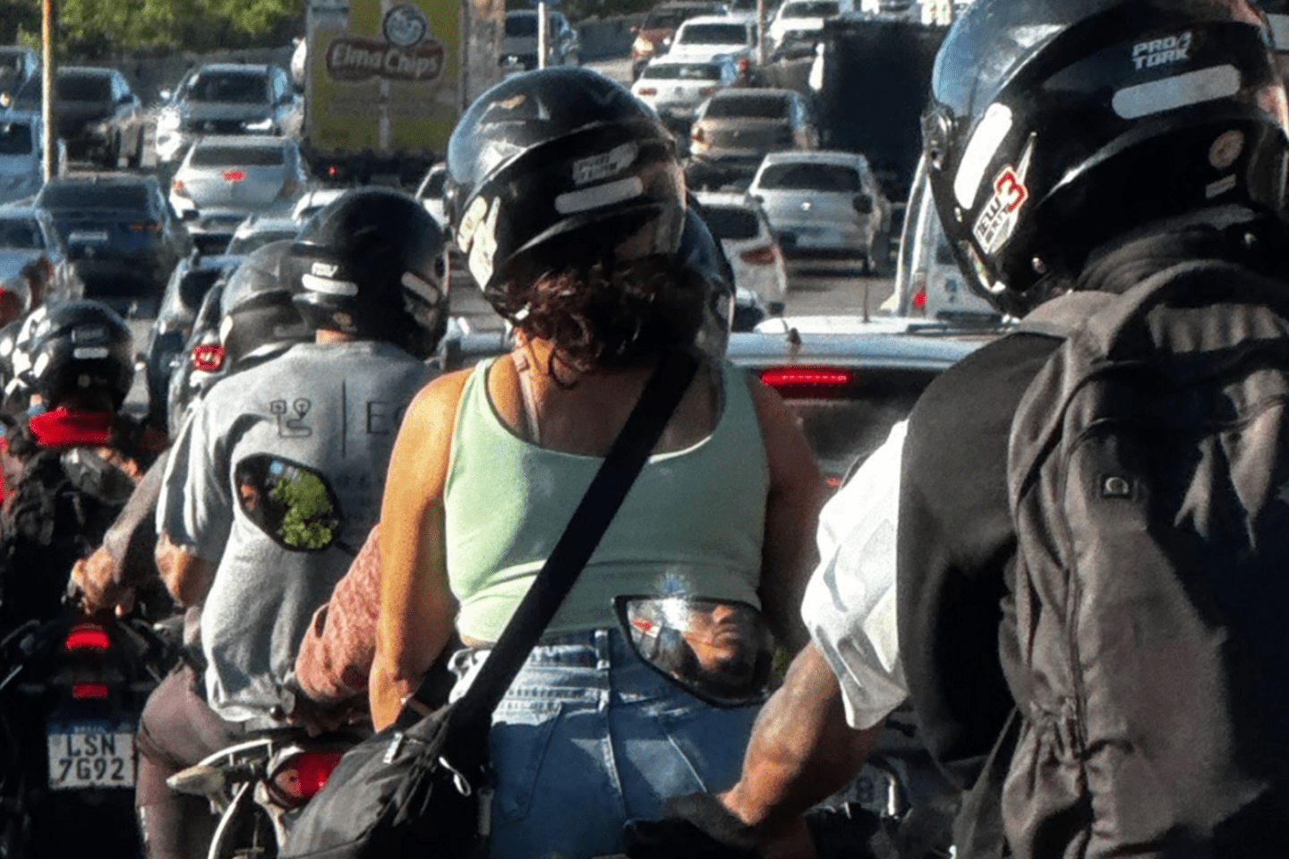 Motociclistas, algunos con pasajeros, circulan entre automóviles en una vía urbana congestionada.
