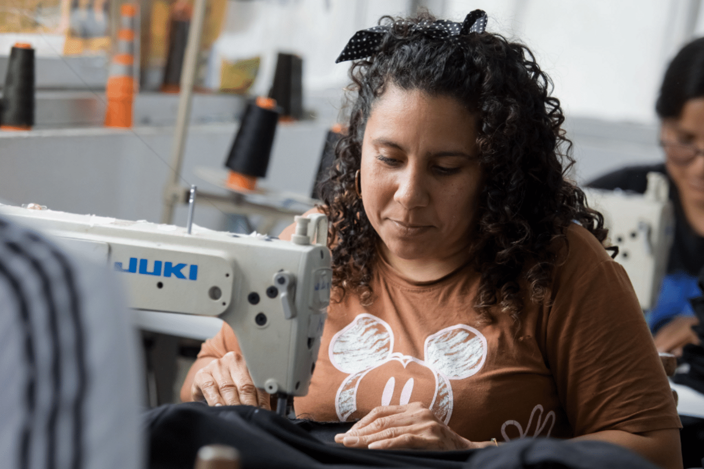 Mujer trabajando en una máquina de coser con otras personas haciendo lo mismo de fondo. 