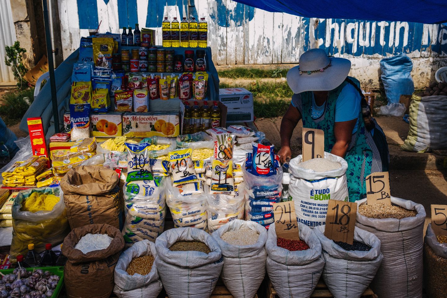 Woman in a wide-brimmed hat arranges products at an open-air market stall, with sacks of grains and cereals and packaged food items on display, prices handwritten on cardboard signs.