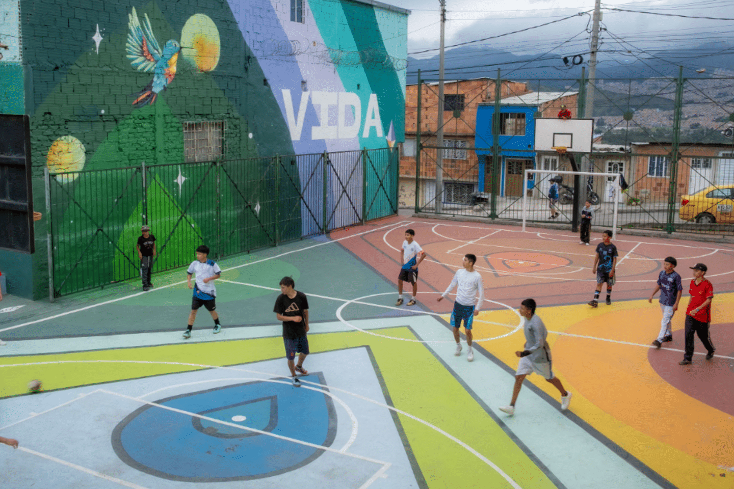 Children playing soccer on a colorful community sports court in Bogotá, Colombia, surrounded by vibrant murals in an urban public space.