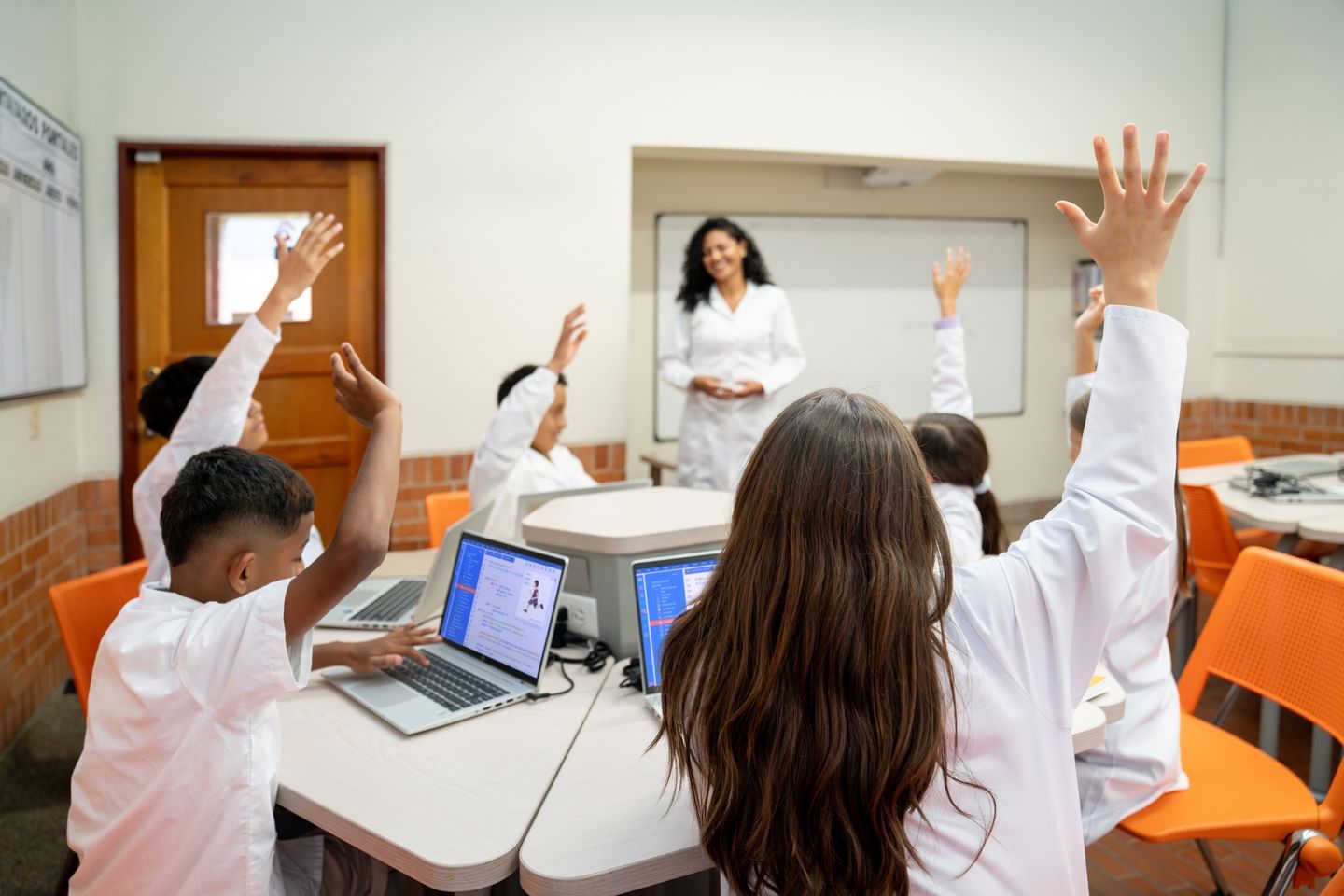Students raise their hands during a classroom lesson while working on laptops, as a teacher stands at the front of the room.