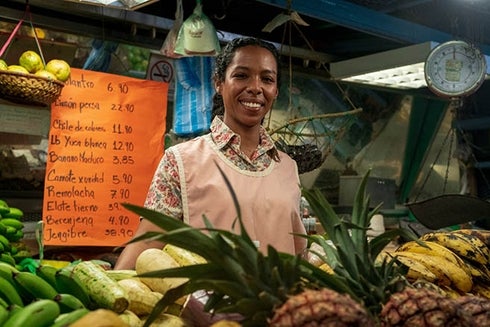A woman standing behind a fruit stand. Social investment - Inter-American Development Bank - IDB