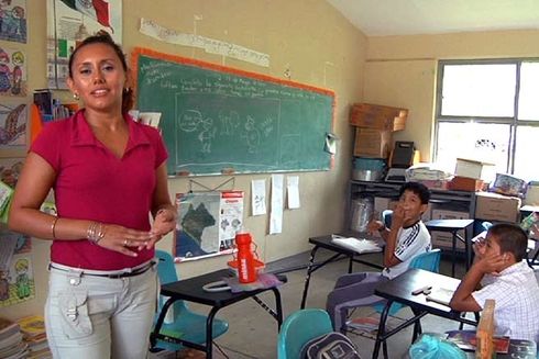 A person standing in a classroom with children in the background. Research - Inter-American Development Bank - IDB