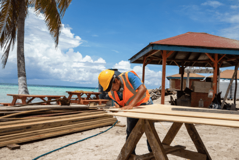 Construction worker in safety gear measuring wooden planks on a tropical beach, with a gazebo and palm trees in the background - Inter-American Development Bank - Development Impact