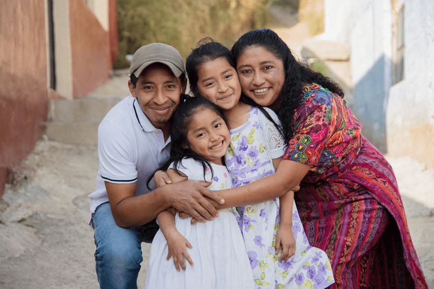 Four members of a migrant family hugging each other with a street in the background - Inter American Development Bank - Migration